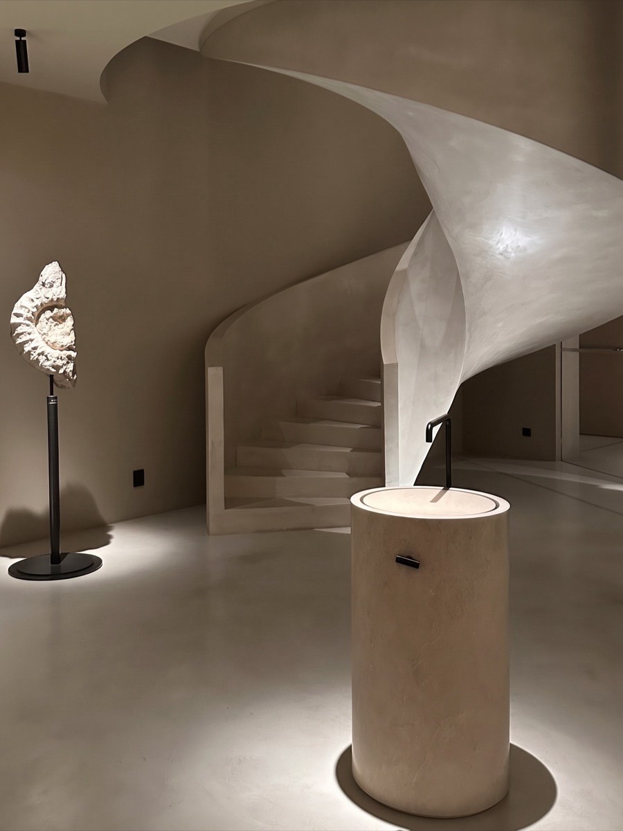 Un escalier de plâtre en spirale, un coquillage sur socle, un fût de pierre éclairé.